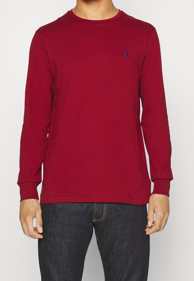 Röd långärmad t-shirt i bomull med rund hals och åtsittande ärmar. Har en liten navy-logotyp på bröstet. Kombinerad med mörka jeans.