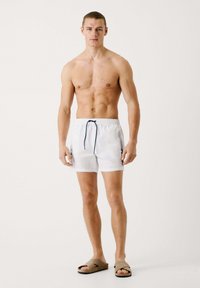 Bañadores blancos con cinturilla elástica y cordón, con acentos en azul oscuro. El modelo está descalzo, llevando sandalias de tela color marrón.
