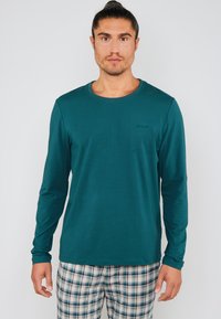 Langærmet skjorte i teal med rund halsudskæring og et lille præget logo. Sammen med ternede bukser i blå, hvid og grønne nuancer.