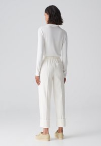 Femme aux cheveux courts et foncés, portant un haut à manches longues blanc, un pantalon large taille haute blanc, et des chaussures beiges, se tenant de dos.