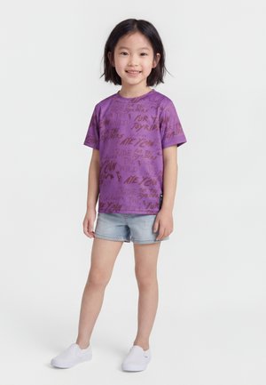 Νεαρή κοπέλα που στέκεται και χαμογελά, φορώντας μοβ μπλουζάκι με σχέδιο, ανοιχτόχρωμα τζιν σορτς και λευκά slip-on παπούτσια σε λευκό φόντο.