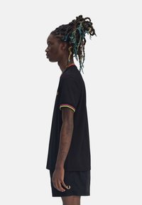 La camiseta polo negra presenta ribetes de colores rojo, amarillo y verde en el cuello y las mangas, combinada con pantalones cortos negros. Detalles del logo dorado visibles.