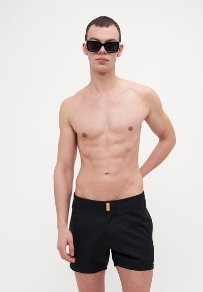 Bañador negro con un diseño ajustado, que presenta un acento dorado en la cinturilla. La tela parece suave y ligera.
