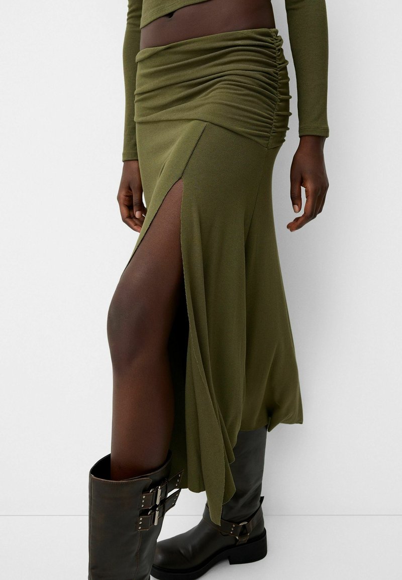 Modèle portant une jupe froncée olive avec une fente haute et des bottes noires à boucle, se tenant devant un fond blanc.