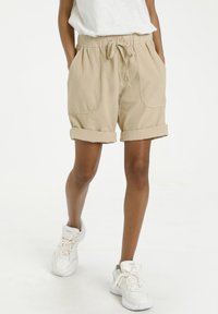 Lichtbeige, knielange shorts met een taillekoord en opgerolde zoom, voorzien van steekzakken aan de voorkant en een textuurstof. Gecombineerd met witte sneakers.