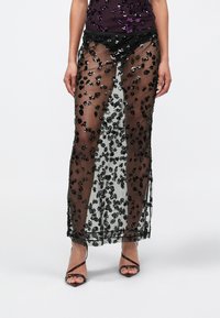 Vrouw die een doorschijnende zwarte rok draagt met glinsterende bloemenpatronen over een zwarte short, gecombineerd met zwarte sandalen met hoge hakken en bandjes.