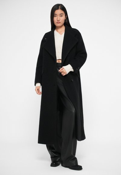 Melnā garā jaka ar izdobi ap kaklu, pogu aizdari un sānu kabatām. Valkāta virs krēmkrāsas īsā adītas džempera un melnām ādas biksēm.