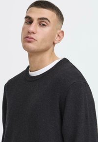 Jeune homme aux cheveux courts portant un pull noir à col rond par-dessus une chemise blanche, regardant légèrement sur le côté devant un fond uni.