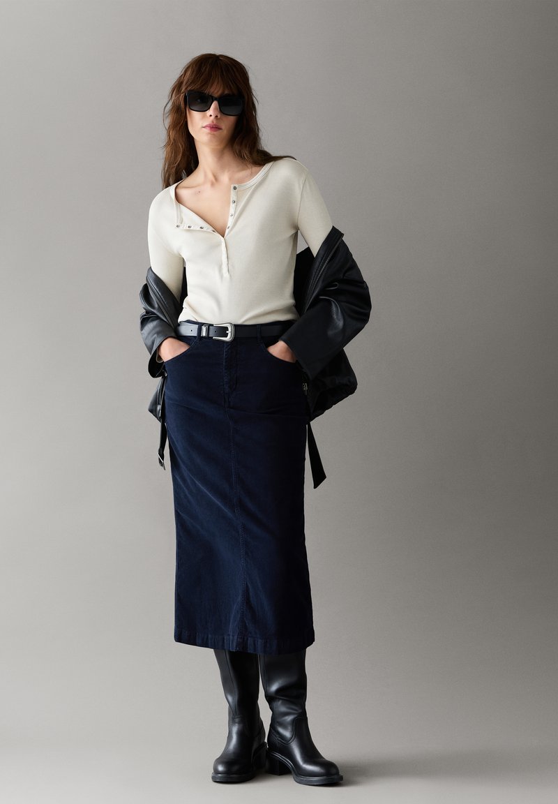 Vrouw met zonnebril, wit topje met knopen, zwart leren jack los over de schouders gehangen, donkerblauwe rok, zwarte riem en zwarte kniehoge laarzen.