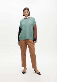 Světle zelený oversized svetr s rukávy v černé, hnědé a tyrkysové barvě, doplněný béžovými kalhotami s rovnými nohavicemi a černými lodičkami se špičatou špičkou.