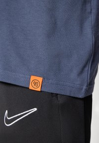 Mørkeblå t-skjorte med rund hals og korte ermer, med oransje merke som har tallet "90". Kombinert med svarte shorts som viser hvit Nike-logo.