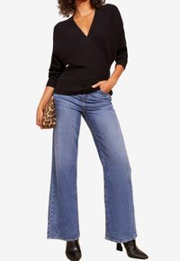 Maglione avvolgente nero a coste con scollatura a V profonda, abbinato a jeans a gamba larga blu chiaro e una pochette con stampa leopardata. Stivaletti neri con punta a stiletto.