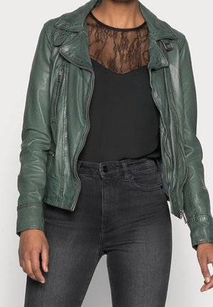 Nainen, joka käyttää tummanvihreää nahkatakkia mustan pitsikauluksisen paidan päällä ja yhdistää sen tummanharmaisiin korkeavyötäröisiin farkkuihin.