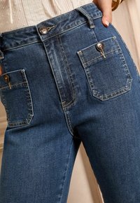 Temno modri kavbojke z visokim pasom, dvema velikima sprednjima žepoma z gumbi in kontrastnim šivom. Na splošno gladka tekstura.