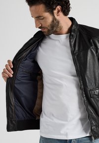 Schwarze Lederjacke mit glatter Textur, Reißverschluss und geripptem Kragen. Das Innenfutter ist navyblau und braun und verfügt über eine Tasche.