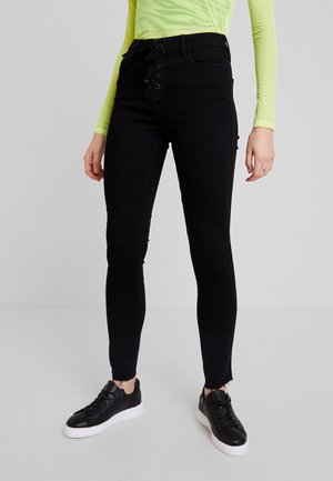 Osoba ubrana w czarne, wysokie, obcisłe jeansy z wiązaniem z przodu, w połączeniu z czarnymi sneakersami i neonowo zieloną bluzką z długim rękawem.