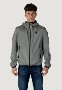 Veste légère grise avec capuche, fermeture éclair frontale et deux poches latérales. Présente des poignets élastiques et un ourlet noir doublé. Tissu texturé.