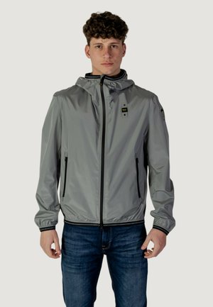 Chaqueta ligera gris con capucha, cremallera frontal y dos bolsillos laterales. Cuenta con puños elásticos y un dobladillo forrado en negro. Tejido texturizado.
