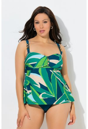 Frau trägt ein Badeoberteil mit grünem, blauem und beige gemustertem Blattmuster mit goldenen Akzenten und passende Bikinihöschen, steht vor einem weißen Hintergrund.