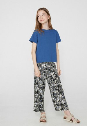 Jeune fille portant un t-shirt bleu, un pantalon large imprimé zèbre et des sandales beiges, debout devant un fond clair uni.