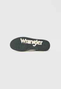 Crna gumena potplat s teksturiranim uzorkom i riječju "Wrangler" istaknutom krem bojom.