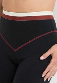 Leggings neri con una fascia bianca e bordeaux, caratterizzati da cuciture bordeaux a contrasto che formano una forma a V in vita.