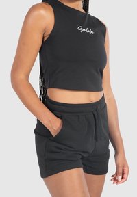 Donna che indossa un top corto senza maniche nero con la scritta "Smilodox" e pantaloni corti neri con coulisse abbinati e tasche, con una mano nella tasca.