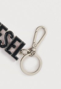 Kulcstartó, amelyen vastag fekete zománcbetűk formázzák a "DIESEL" feliratot, ezüst fém csipesszel és kör alakú kulcskarikával rögzítve. Sima, fényes felület.