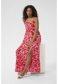 Robe longue rouge et blanche à fleurs avec un corsage ajusté et une jupe fluide dotée d'une fente latérale. Le modèle porte des sandales blanches à lanières.