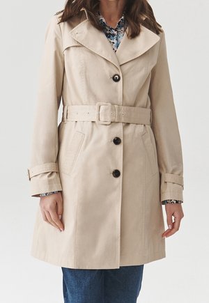 Trench-coat beige longueur genou avec fermeture à boutons, ceinture à la taille et poches latérales, porté sur un chemisier fleuri et un jean bleu.