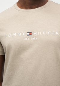 Μπεζ βαμβακερό t-shirt με λευκό κέντημα που γράφει "TOMMY HILFIGER EST. 1985" και μικρό λογότυπο σε κόκκινο, λευκό και μπλε πάνω από το όνομα της μάρκας.