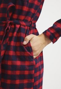 Czerwona i granatowa koszula flanelowa w kratę z przewiązanym w pasie i boczną kieszenią, z widoczną fakturą tkaniny i długim rękawem.