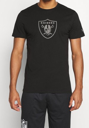 Mężczyzna ubrany w czarną koszulkę Raiders z logo przedstawiającym pirata z opaską na oku i skrzyżowanymi mieczami, w połączeniu z czarnymi szortami NFL.