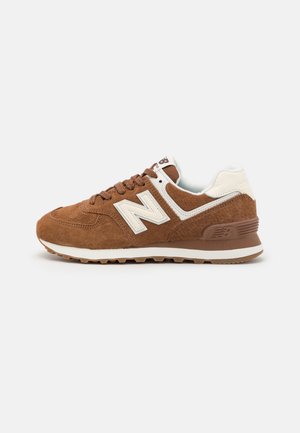 Brun ruskind New Balance-sneaker med hvide detaljer, tekstureret hæl og gummisål, set fra ydersiden på en hvid baggrund.