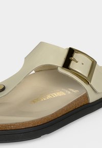 Μπεζ σανδάλι με καμπυλωτό φελλό πάτο, μαύρη σόλα, ρυθμιζόμενο χρυσό λουράκι με πόρπη και δαχτυλίδι στον ανοιχτό γκρίζο φόντο.