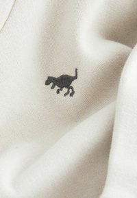 Tessuto crema con una superficie liscia caratterizzato da un piccolo design di un gatto nero ricamato. Il materiale è morbido e leggermente elastico.