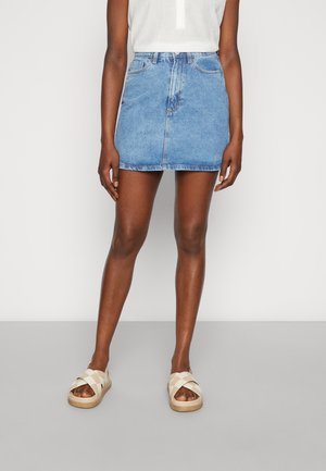 Osoba ubrana w niebieską dżinsową spódnicę sięgającą do połowy uda, biały bezrękawnik oraz beżowe sandały z krzyżującymi się paskami stojąca na szarej podłodze.