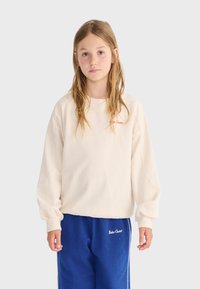 Joven con cabello largo que lleva una sudadera holgada color crema y pantalones deportivos azules con el logo "Bobo Choses" en ambas prendas.