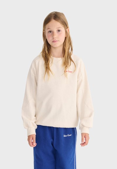 Jeune personne aux longs cheveux portant un sweat-shirt oversize crème et un pantalon de survêtement bleu avec le logo « Bobo Choses » sur les deux vêtements.