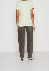 Mentolově zelené tričko s krátkým rukávem v kombinaci s volnými hnědými kalhotami s vysokým pasem a tělovými podpatky, stojí před jednobarevným pozadím.