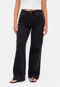 Crne traperice širokih nogavica s visokim strukom, izrađene od trapera. Imaju prednje džepove, zatvaranje na gumb i čist završetak. Tkanina s teksturom.