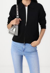 Černý zip-up kardigan s límečkem, vyrobený z měkké látky. Kombinován s světlými modrými džíny a malou stříbrnou kabelkou.