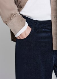 Dunkelblaue Cordhose mit strukturierter Oberfläche, kombiniert mit einem beigen Hemd mit Knopfleiste und weißen Manschetten. Sichtbare Hand in der Hosentasche.