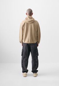 Beige Kapuzenpullover mit lockerer Passform. Dunkelgraue Cargo-Hosen mit mehreren Taschen. Beigefarbene Schuhe. Neutraler Hintergrund.