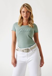 T-shirt verde attillato con logo "Guess" in filo metallico, abbinato a jeans bianchi a gamba larga e una cintura bianca con borchie. Tessuto morbido e liscio.