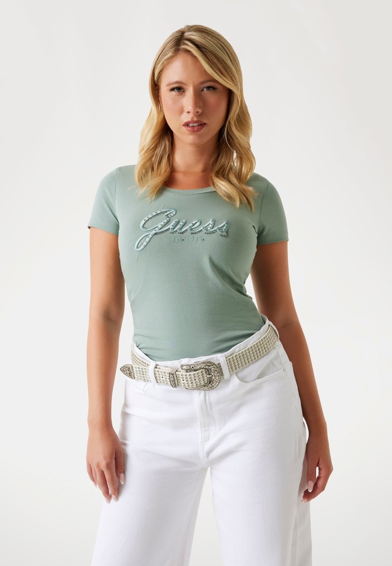 Green fitted t-shirt with "Guess" logo in metallic thread, paired with white wide-leg jeans and a studded white belt. Smooth, soft fabric.