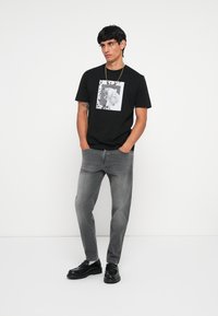 Čierne tričko s okrúhlym výstrihom a predným grafickým potlačením, v kombinácii so sivými džínsy a čiernymi slip-on topánkami. Hladké textúry a voľný strih.