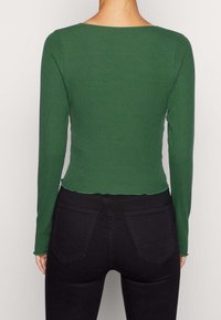 Grünes geripptes Langarmshirt mit rundem Halsausschnitt und gewelltem Saum, kombiniert mit schwarzen Jeans mit normalen Gesäßtaschen.