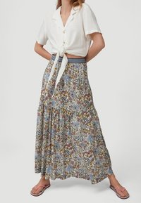 Femme portant un chemisier blanc à manches courtes noué, une jupe maxi à motifs floraux avec une ceinture rayée, et des tongs roses, debout les jambes écartées.