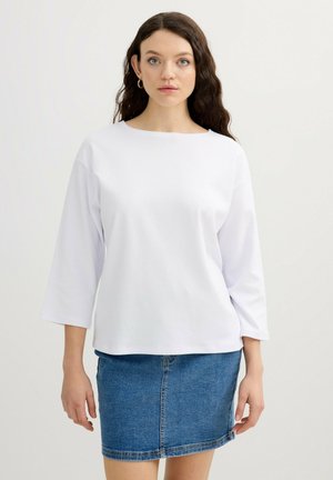 Mujer con cabello largo y rizado que lleva pendientes de aro dorados, camiseta blanca de manga larga y falda vaquera azul sobre un fondo liso.